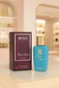 A perfume bottle and its box are displayed on a reflective surface in front of a blurred background of shelves with various items. Produktbild-Vorschau