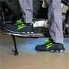 A person wearing black and green safety shoes is standing next to a motorcycle. Voir les images du produit