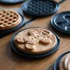 A close-up shot of several waffle plates on a wooden table. One plate has a gingerbread-shaped waffle on it, and another has a round waffle. Náhled obrázku produktu