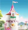 A toy princess castle with a princess figure in a window, set against a backdrop of rolling hills and a blue sky. Voir les images du produit