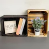 Two wooden crates are displayed side-by-side against a textured wall. The left crate is dark, containing a square sign with the word 'HOME' and two books. The right crate is lighter wood and holds a potted plant with light blue flowers. Produktbild-Vorschau