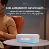 A man sits in a chair next to a table with a digital alarm clock on it. The alarm clock has a digital display showing the time and temperature. Produktbild-Vorschau