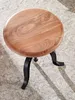 A round wooden stool with black metal legs sits on a patterned rug. Produktbild-Vorschau