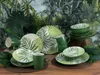 A collection of green and white patterned tableware, including plates, bowls, and mugs, arranged on a wooden surface with a tropical jungle-themed wallpaper in the background. Náhľad obrázku produktu