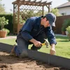 A man is kneeling in a garden, hammering stakes into the ground to install a black garden edging. Produktbild-Vorschau