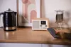 A kitchen counter scene with a kettle, a digital radio, and a person holding a smartphone. Voir les images du produit