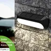 A solar-powered wall lamp is mounted on a stone wall, with a blurred background of a lake and hills. Produktbild-Vorschau