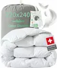 A folded white duvet is presented in front of a gray storage bag with product information printed on it. Several white feathers are scattered around the image, and a Swiss flag tag is attached to the duvet. Produktbild-Vorschau