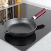 A cast iron frying pan with a wooden handle sits on a black glass stovetop. Jars of pasta and other dry goods are visible in the background. Náhľad obrázku produktu