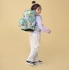 A young girl wearing a colorful backpack and striped pants stands against a plain yellow background. Podgląd zdjęcia produktu