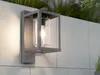An outdoor wall lamp is mounted on a tiled wall, with plants in the background. Anteprima dell’immagine prodotto