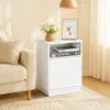 A white nightstand with two drawers and an open shelf is placed next to a cream-colored sofa in a room with light-colored walls and curtains. Náhled obrázku produktu