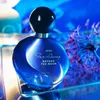 A blue perfume bottle with a black cap, placed on a reflective surface with a blurred blue and purple background. Náhľad obrázku produktu