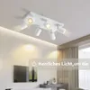 A white ceiling light fixture with multiple adjustable spotlights is shown mounted on a white ceiling. The room below is softly lit, with a television and furniture visible in the background. Produktbild-Vorschau
