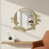 A round mirror with a decorative gold frame featuring leaf motifs is mounted on a light-colored wall above a wooden shelf. A small green vase with a sprig of dried leaves sits on the shelf next to a stack of books. Part of an orange chair is visible in the Voir les images du produit