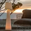 A floor lamp is placed on a wooden deck next to a chair, with a sunset over the ocean in the background. Produktbild-Vorschau