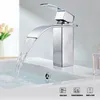 A modern chrome waterfall faucet is shown in operation over a white sink, with a vase of flowers in the background. Produktbild-Vorschau