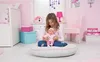 A young girl is sitting on the floor in a pink-themed bedroom, holding a baby doll. Náhled obrázku produktu
