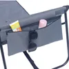 A close-up view of a gray folding chair with a built-in storage pocket. The pocket contains a tube of sunscreen, a pair of sunglasses, and a pink bottle. Produktbild-Vorschau
