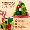 A hand is placing a small decorative box onto a tiered wooden advent calendar shaped like a Christmas tree. The background is a light yellow with snowflake graphics. Produktbild-Vorschau