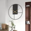 A modern wall clock is displayed on a white wall, with a bookshelf and part of a sofa visible in the background. Produktbild-Vorschau