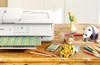 A Canon PIXMA TS9551Ca printer is shown on a wooden desk, printing a patterned sheet of paper. Various craft supplies and decorative items, including paper flowers, pencils, a chipmunk figurine, a panda figurine, and a parakeet figurine, are arranged aroun Podgląd zdjęcia produktu