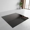 A black glass hearth plate is placed on a concrete floor in a room with a white wall and a window. Produktbild-Vorschau