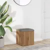 A cube-shaped storage ottoman with a wood-grain finish and a gray upholstered top sits on a wooden floor next to a large potted plant. A white wall and a partial view of a door are in the background. Anteprima dell’immagine prodotto