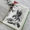 An open book with an illustration of an eagle and a small animal, with a dried flower as a bookmark. Produktbild-Vorschau