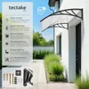 A tectake canopy is mounted above a door on a white house, with green plants in the foreground. Náhled obrázku produktu