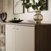 A close-up view of a modern sideboard with two doors, showcasing its textured wooden side and a beige front. Decorative items like a black bowl with greenery and a glass vase with eucalyptus leaves are placed on top. Produktbild-Vorschau