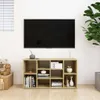 A television is mounted on a wall above a light wood media console filled with decorative items and books. The console has multiple cubbies and a plant is visible to the right. Podgląd zdjęcia produktu