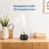 An aroma diffuser is shown on a wooden dresser, emitting steam. Decorative elements like a vase with plants and books are also present. Produktbild-Vorschau