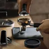 A person is using a coffee tamper to press coffee grounds in a portafilter, with other coffee accessories laid out on a wooden table. Anteprima dell’immagine prodotto