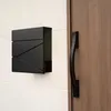 A black mailbox mounted on a white wall next to a wooden door with a black handle. Produktbild-Vorschau