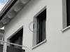 A close-up view of a white building exterior, showing a section of the wall with a window and a small, dark rectangular element highlighted by a blue circle. Produktbild-Vorschau