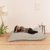 A woman is lying on a large, light gray floor cushion in a room with a lamp and a small table. Voir les images du produit