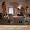 Two women are using cordless vacuum cleaners in a modern, well-lit living room with a dog sitting on a chair. Produktbild-Vorschau