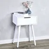 A white bedside table with a drawer and three legs, placed against a light gray wall. A book and a vase with decorative branches are on top of the table. Produktbild-Vorschau