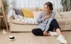 A woman is sitting on the floor in a living room, looking at a small robot. Anteprima dell’immagine prodotto