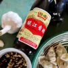 A bottle of Zhenjiang vinegar is shown next to a head of garlic and a bowl of dipping sauce, with dumplings in a green and white bowl in the foreground. Podgląd zdjęcia produktu