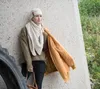 A woman leans against a concrete wall, holding a jacket and a bag, with a large tire to her left and greenery in the background. Produktbild-Vorschau