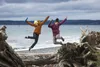 Two people jumping on a beach with driftwood in the foreground and the ocean in the background. Produktbild-Vorschau
