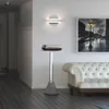A modern living room with a white tufted sofa, a black and white framed picture of a street scene, and a minimalist floor lamp with a turntable on top. Produktbild-Vorschau