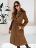A woman wearing a brown winter coat and sunglasses stands in front of a large potted plant. Náhled obrázku produktu