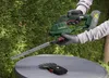 A person is using a cordless grass and shrub shear in a garden setting. Produktbild-Vorschau