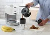 A person is pouring coffee from a carafe into a travel mug next to a coffee maker, with bananas and granola bars on the counter. Voir les images du produit