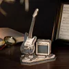 A wooden model of an electric guitar and amplifier on a wooden surface, with a book and magnifying glass in the background. Voir les images du produit