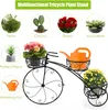 A black metal tricycle-shaped plant stand with three baskets, holding potted plants and a watering can. The top of the image displays various types of plants with labels. Produktbild-Vorschau