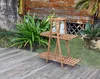 A wooden plant stand is placed on a wooden deck in front of a wooden wall and some greenery. Produktbild-Vorschau